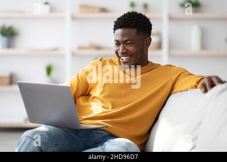 Joyful black guy sitting on couch with laptop Stock Photo