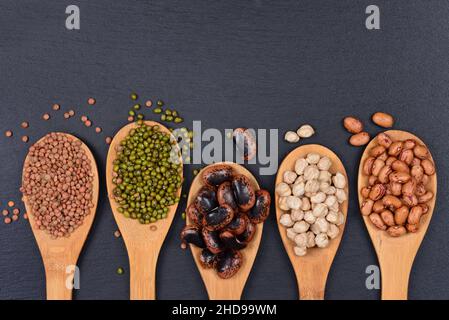 Various legumes lie on wooden spoons and are photographed from above on a dark slate of slate Stock Photo