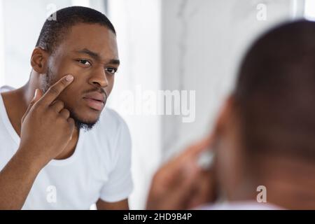 Concerned Black Young Man Examining His Face While Looking At Mirror Stock Photo