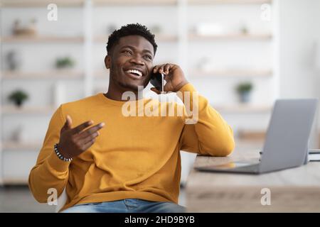 Smiling man sitting at table with laptop, having phone conversation Stock Photo