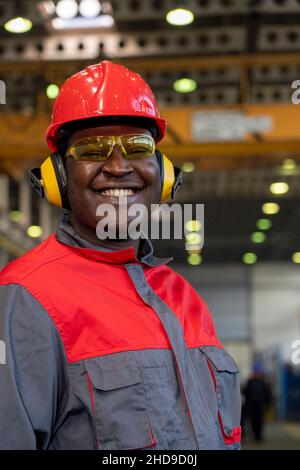 Smiling Black Worker In Personal Protective Equipment Looking At Camera. Portrait Of Industrial Worker In Red Helmet, Safety Goggles And Work Uniform. Stock Photo