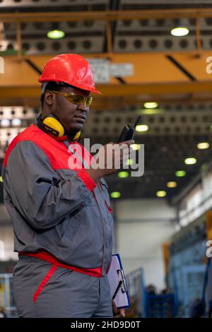 African American Worker In Protective Workwear Talking On Radio Communication Equipment In A Factory. Portrait Of Black Industrial Worker. Stock Photo