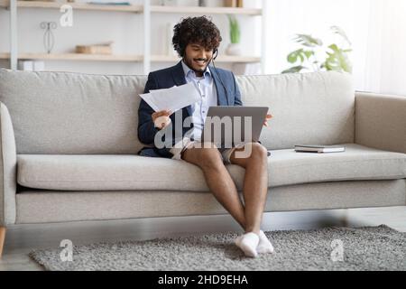 Attractive indian guy sitting on couch, using laptop and headset Stock Photo