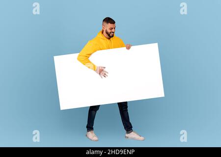 Excited arab man walking with white blank placard under arm over blue studio background, mockup for text or design Stock Photo