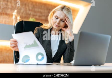 Serious successful pensive smart caucasian mature business lady, financial analyst, manager, sitting at her desk in office, studying financial charts, analyzing and planning financial strategy Stock Photo