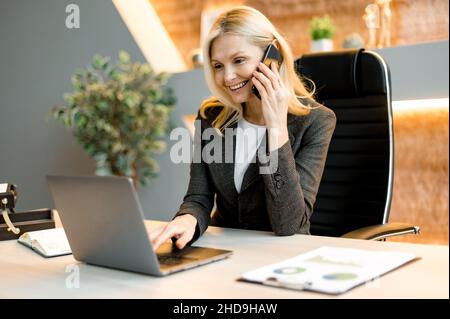Mature happy joyful confident caucasian blonde business lady, broker or manager, sitting at table in a modern office, working with laptop, talking to client or employees on smartphone, smiling Stock Photo