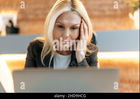 Smart focused exhausted mature woman, business woman or broker, in formal suit, tired of office work, experiencing stress and headache, massaging her temple, needs rest, looking languidly at screen Stock Photo