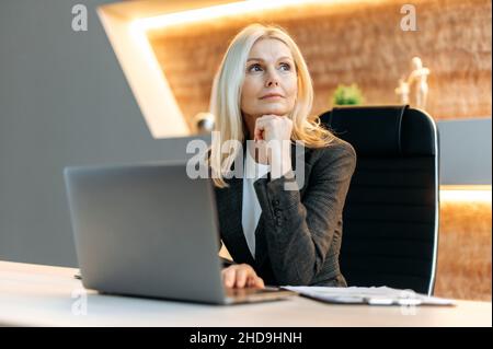 Influential successful smart blonde caucasian mature business woman, financial analyst, freelancer, sitting at table, taking break from work, looking thoughtfully aside, dreaming about vacation Stock Photo