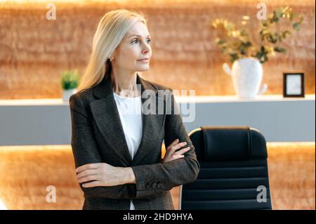 Portrait of serious influential beautiful caucasian blonde middle aged business woman, ceo of the company, stand in a modern office in a formal suit, pensively looking to the side with crossed arms Stock Photo