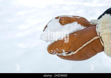 Woman in leather mittens rolls a snowball outdoors, close-up. Hands in warm mittens make snow in winter. Brown leather mittens in the snow in winter. Stock Photo