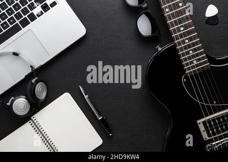 A laptop, headphones and a pen are on a black table next to a guitar, sunglasses and picks. Creative youth background concept. Stock Photo