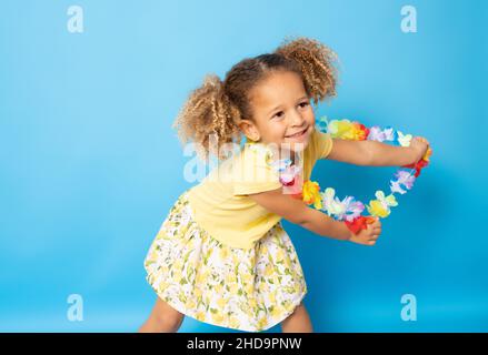 Happy little girl wearing Hawaiian necklace dancing isolated over blue background. Stock Photo