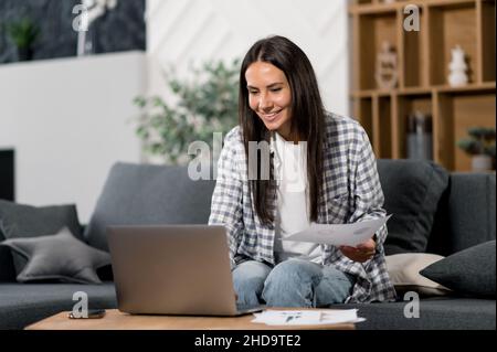 Caucasian beautiful positive young woman, in casual wear, freelancer, working remotely at home, using laptop, checking documents, planning, developing strategy, in living room on sofa. Distant work Stock Photo