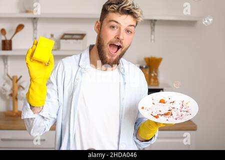 Surprised young man with sponge and dirty plate in kitchen Stock Photo