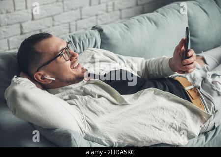 Man wearing eyeglasses holding mobile phone and smiling while lying on sofa. Airpods. Wireless communication. Mobile communication. Stock Photo