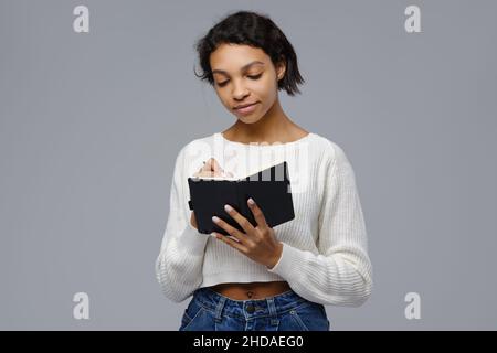 Concentrated young african woman standing against grey background and writing in diary Stock Photo
