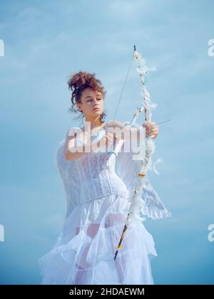 Valentines Day. Love and romance. Teenager angel girl with wings aiming with bow and arrow, copy space on sky. Stock Photo