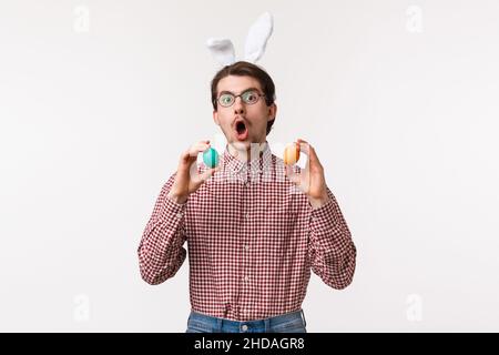 Traditions, religious holidays, celebration concept. Funny joyful young man with beard in glasses, wear cute rabbit ears and hold two painted eggs Stock Photo