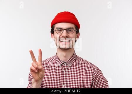 Close-up portrait of cute happy smiling caucasian guy, showing peace gesture and looking camera friendly, informal greeting, wearing hipster beanie Stock Photo
