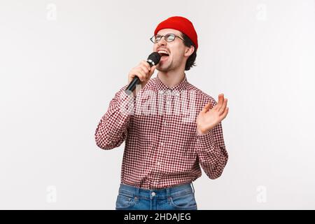 Brokenhearted guy singing romantic song in karaoke with passionate feeling, close eyes and scream into microphone, standing in red beanie, glasses and Stock Photo