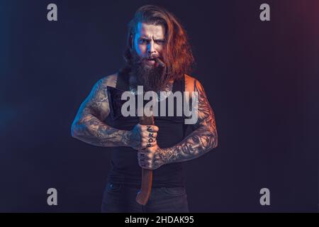 Handsome man holds old axe. Bearded lumberjack. Serious male with tattoos with long hair. Stock Photo