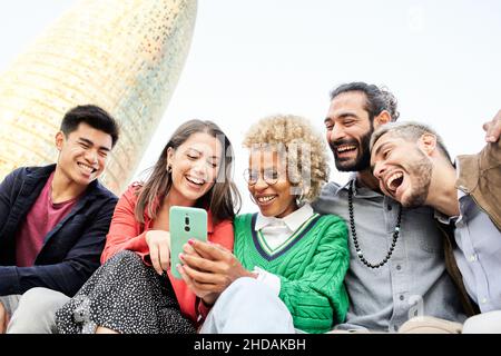 Group of multi ethnic friends in college together outdoors enjoying watching text or video on mobile phone. Smiling group of students Stock Photo