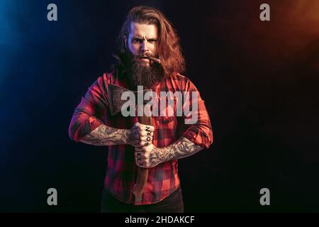 Handsome man with old axe. Bearded lumberjack. Serious brutal male in checkered shirt with long hair Stock Photo