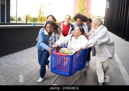 Crazy friends making a racing challenge with shopping cart. Multiracial group of young people having fun outside Stock Photo