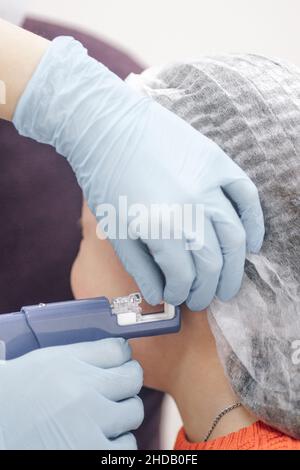 Close-up of nurse hands in disposable medical gloves pierce the ear with gun for piercing ears of child. Process of piercing ears of little girl. Stock Photo