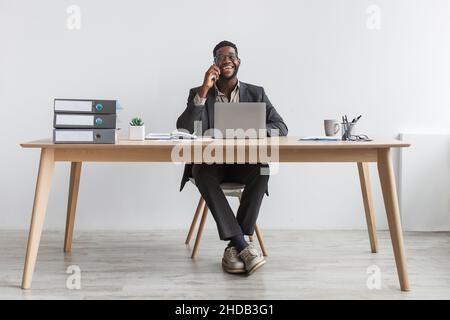 Cheerful black businessman in formal suit talking on smartphone, having conversation, sitting at table with laptop Stock Photo