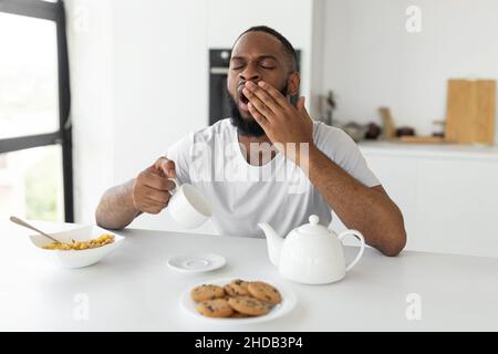 Black man yawning, pouring coffee away from cup Stock Photo