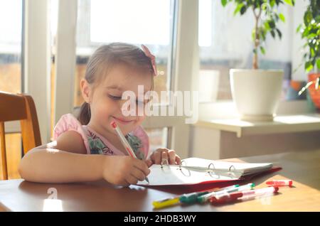 Adorable smiling little girl sitting at table and drawing with multicolor gel pens in notebook, happy kid enjoying art activity during leisure time ho Stock Photo