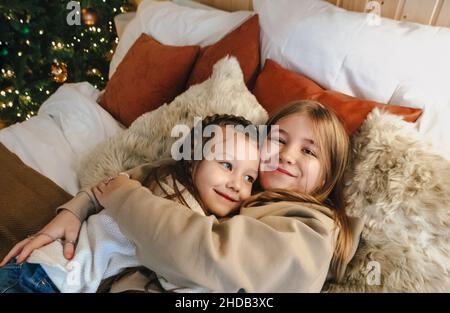 Cozy domestic portrait of two happy smiling little girls sisters hugging embracing while spending leisure time together at home, lying on bed near dec Stock Photo