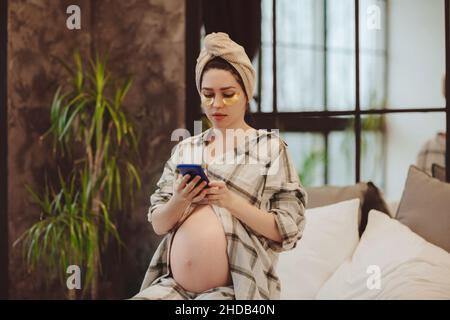 Beauty and skin care during pregnancy. Young pregnant woman in pajama with head wrapped in towel with cosmetic patches under eyes using mobile phone w Stock Photo