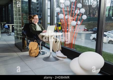 Redhead freelancer using laptop near backpack in hotel lobby Stock Photo