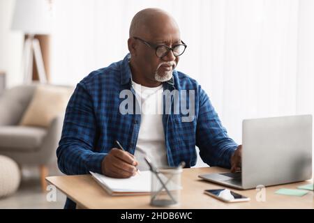Black Man Using Laptop Taking Notes In Office, Wearing Eyeglasses Stock Photo
