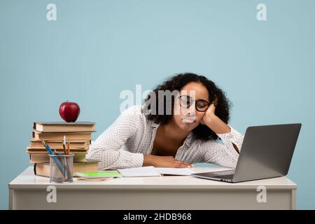 Education and overworking. Tired black female student studying at desk with laptop against blue studio background Stock Photo