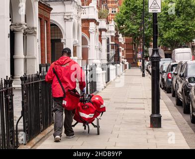A postman (mail delivery) delivering mail/post with a wheeled trolley along Harrington Gardens, Kensington, London Stock Photo