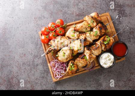 Juicy grilled chicken skewers with fresh vegetables and two sauces close-up on a wooden tray on a gray concrete background. horizontal top view from a Stock Photo