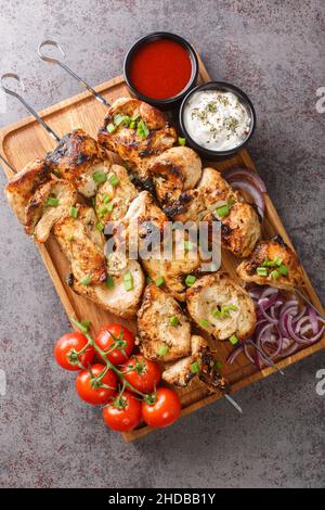 Homemade chicken kebab on skewers with vegetables and two sauces close-up in a wooden board on gray concrete background. Vertical top view from above Stock Photo