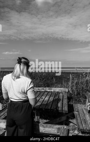 Vertical grayscale shot of a woman standing near a wooden bench on a seaside Stock Photo