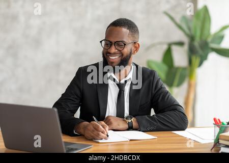 African American man in glasses using laptop writing in notebook Stock Photo