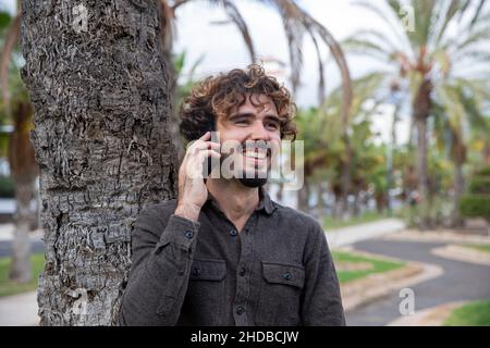 Man makes a phone call and is smiling and happy. Person outside in the fresh air in a park with palm trees. Caucasian man happy to talk on the phone. Stock Photo