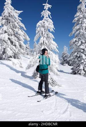 happy woman in a ski suit, helmet, stands in the mountains against the background of ice trees on a sunny winter day. a minute of rest, relaxation, pl Stock Photo