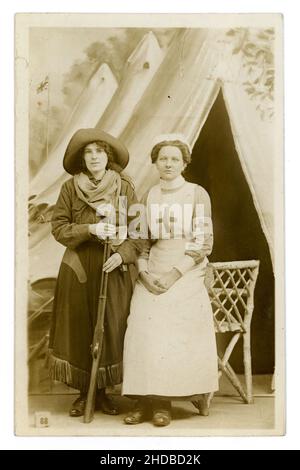 Original WW1 era postcard of 2 attractive young women, friends, having fun, posing for a photo in fancy dress costume one is dressed as cowboy / cowgirl with wide brimmed hat holding a rifle, and the other as the iconic red cross nurse - from a popular London studio called the Fancy Dress Studio at 49 Tottenham Court Road, London  circa 1914, 1915 Stock Photo