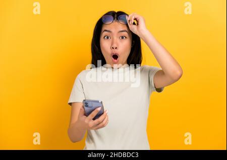 Shocked amazed chinese young brunette woman, holding a cellphone in hand, looks surprised at camera, takes off her glasses opening mouth, standing on isolated orange background in casual t-shirt Stock Photo