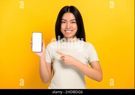 Happy chinese positive girl in casual t-shirt showing modern smart phone with empty white screen and points a finger at him, stands on isolated orange background, smiling. Mockup, copy space Stock Photo