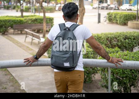 back shot of male tourist student leaning on a railing with a backpack looking into a park Stock Photo
