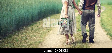 wedding couple, bride and groom holding hands and walks the country road beside cereal field. back view copy space Stock Photo