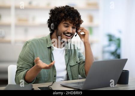 Cheerful indian guy sitting at table, using laptop and headset Stock Photo